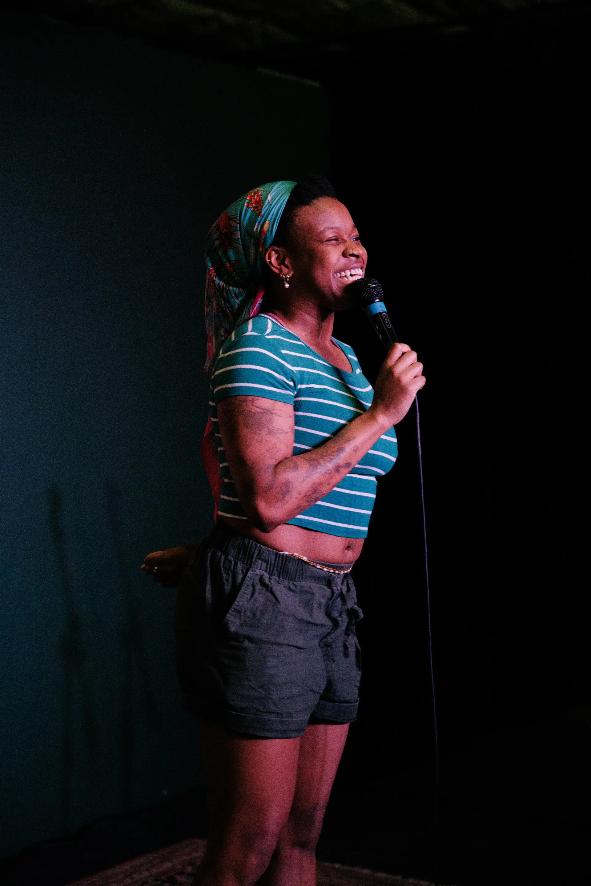Sami Beason laughing into a microphone on a dark stage in a teal striped top and headwrap