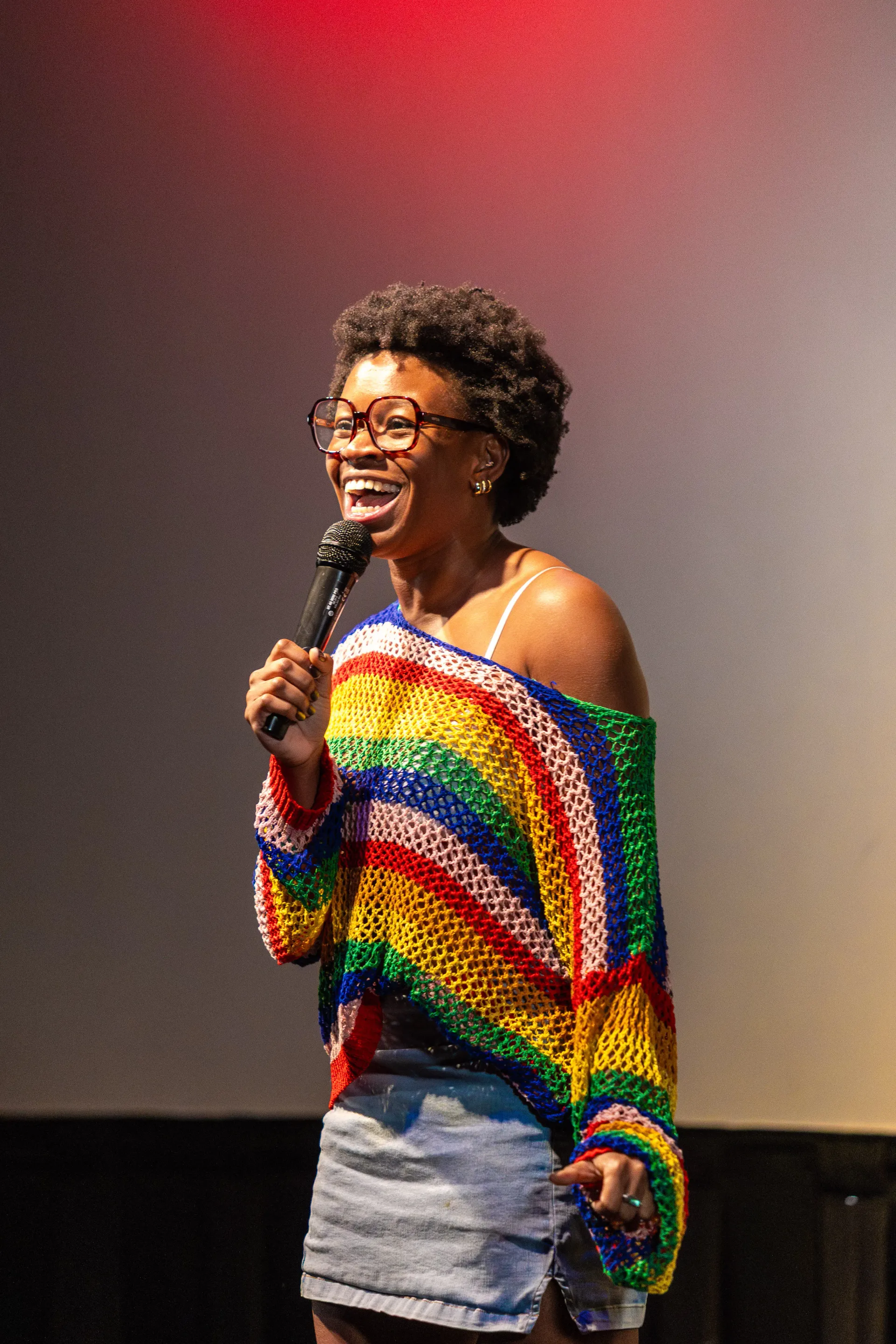 Sami Beason laughing into a microphone in a rainbow crochet sweater onstage