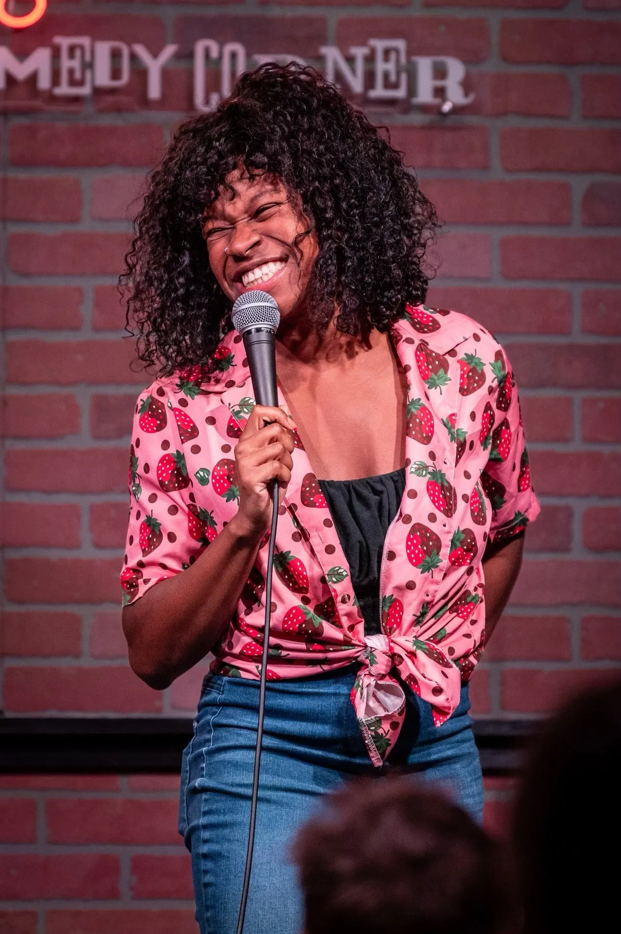 Sami Beason laughing into a microphone in a pink strawberry-print shirt onstage